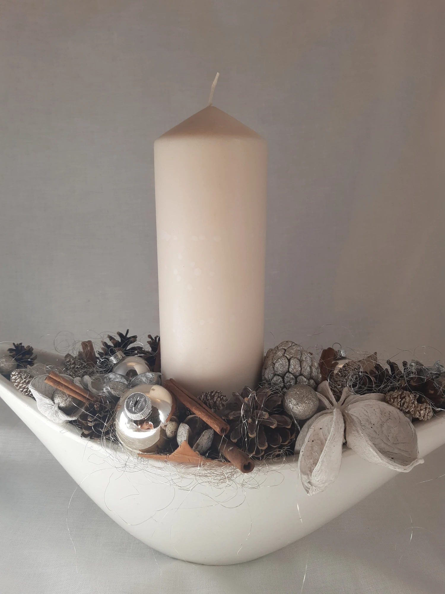 Arrangement décoratif de noël, jolie bougie crème dans un vase en céramique blanche accompagnée d'éléments naturels et de boules de noël.